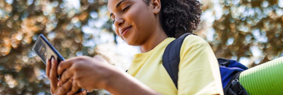 A young back woman outside with a backpack on, looking down at her phone