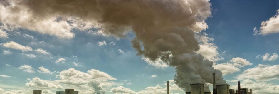 industrial buildings releasing grey smoke into the air, next to crops