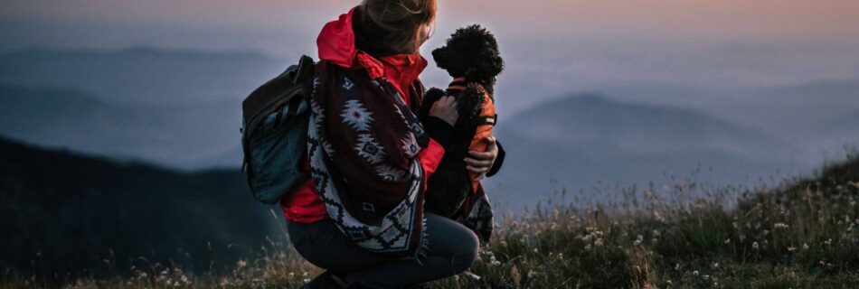 A woman at dusk kneeling with a dog, looking at the sunset
